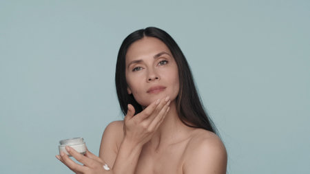 Cream for skin care. A seminude brunette woman applies cream to her face. Woman with a jar of cream in her hands in the studio on a blue background close up.の写真素材