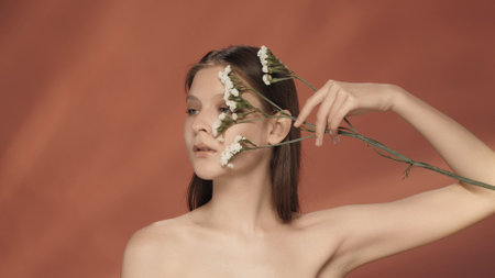 Portrait of a woman with white wildflowers near her face close up. Seminude woman in the studio on a red background. The concept of beauty, cosmetology, treatment and care.の写真素材