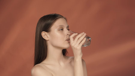 A young woman drinks clean, clear water from a glass. Seminude woman in the studio on a red...