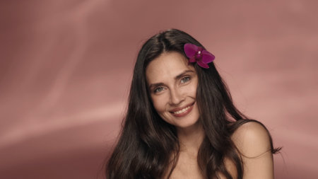 Portrait of a seminude woman with an orchid flower in her hair in the studio on a pink background close up. Cosmetic line for skin, hair care with orchid extract. The concept of beauty, cosmetology, care.の写真素材