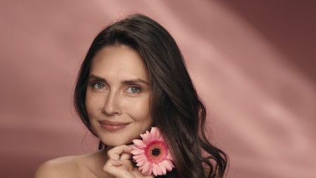 A woman touches a gerbera flower to her perfect skin of face. Portrait of a woman with a gerbera flower in the studio on a pink background close up. The concept of beauty, cosmetology, skin care.の写真素材