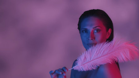 Close up shot of a seminaked young woman covering herself with two feathers in blue and pink color scheme and shadowed background.の写真素材
