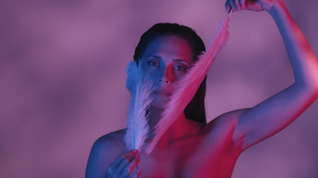 Close up shot of a seminaked young woman touching her face with two feathers in blue and pink color scheme and shadowed background.の写真素材