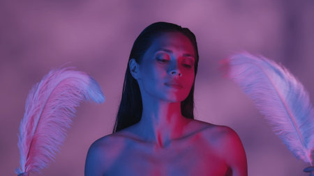 Close up shot of a seminaked young woman waving two feathers next to her face in blue and pink color scheme and shadowed background.の写真素材