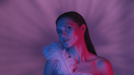 Close up shot of a seminaked young woman covering herself with two feathers in blue and pink color scheme and shadowed background.の写真素材