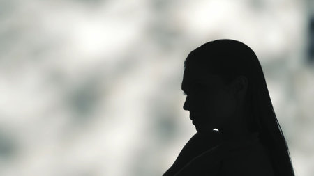 Close up side profile shot of a young womans black silhouette, shes blowing a feather off her palm on a shadowed light green background.の写真素材