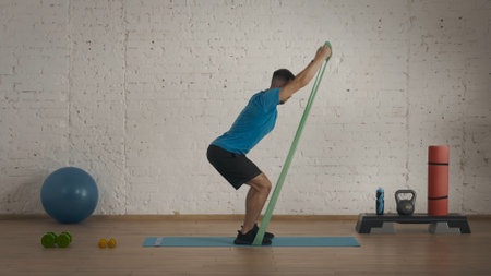 Man fitness instructor in sportswear doing half squat rubber band exercise at the home studio.