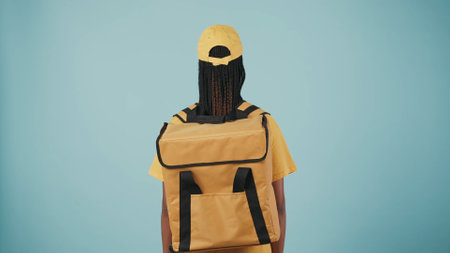 Delivery woman in uniform with portable backpack refrigerator, posing at the camera with her back. Isolated on blue background.の写真素材