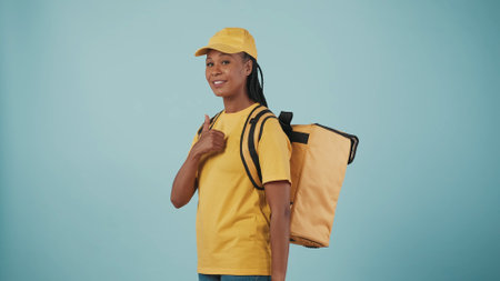 Delivery woman in uniform with portable backpack refrigerator, showing thumbs up gesture and smiles. Isolated on blue background.の写真素材