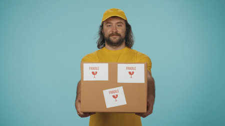 Delivery man in yellow uniform smiling, holding a box with fragile stickers. Isolated on blue background.の写真素材
