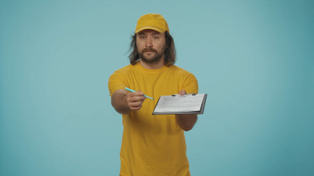 Delivery man in yellow uniform gives the order documents to sign at the camera. Isolated on blue background.の写真素材