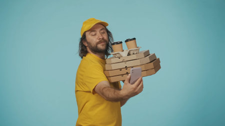 Delivery man in uniform holding pizza boxes and coffee taking a selfie video. Isolated on blue background.の写真素材