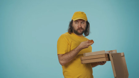 Delivery man in yellow uniform holding stack of pizza boxes, eating the big slice. Isolated on blue background.の写真素材