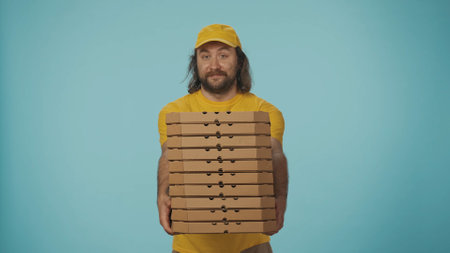 Delivery man in yellow uniform holding a big stack of pizza boxes. Isolated on blue background.