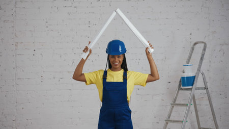 Medium shot of a dark-skinned young female construction worker raising two rolled up plans above his...