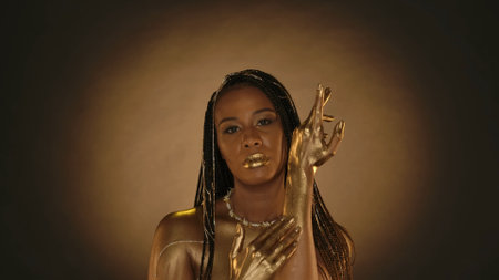 A woman with golden lips, golden skin on a brown background with circular light. A seminude African American woman applies, rubs golden paint, liquid gold into the skin of her hands.の写真素材