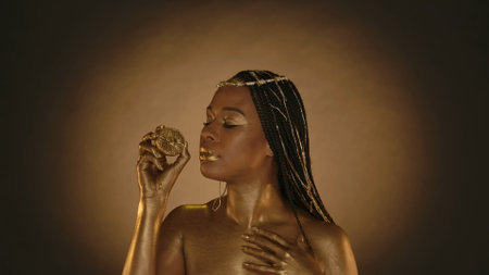 African American woman in Cleopatra style on brown background with circular light. A woman with skin in golden paint, with jewelry on her head, holds a half of a lemon. Fashion art design.の写真素材
