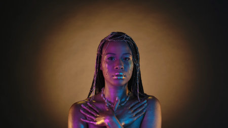 Portrait of a woman with golden drops of tears on her face in pink neon light. African American woman with golden metallic make up, with jewelry in her hair on a brown background with circular light.の写真素材