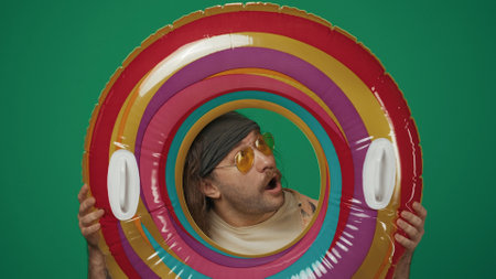 Man in casual clothing holds inflatable tube ring and looking through it at the upper right direction with amazed face expression. Isolated on green background.の写真素材