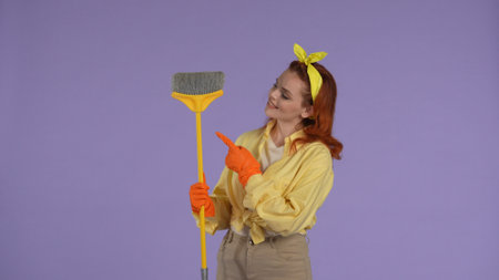 Woman in casual clothing and rubber gloves holding broom pointing ai it, smiling face, looking at...