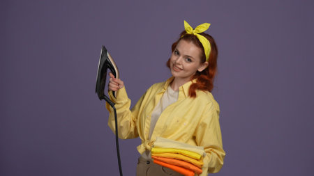 Woman in casual clothing holding stack of folded tshirts and iron, smiling at the camera. Isolated on purple background.の写真素材