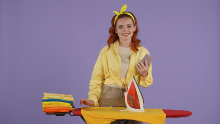 Woman in casual clothing and headband ironing shirts, holding smartphone and looking at the camera. Isolated on purple background.の写真素材