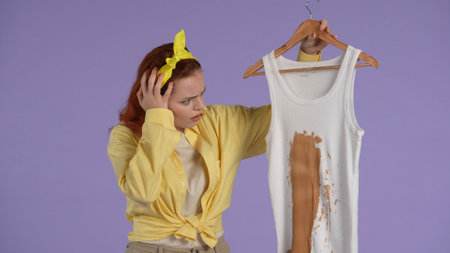 Woman in casual clothing and rubber gloves holding piece of clothing with big brown stain, helpless face. Isolated on purple background.の写真素材