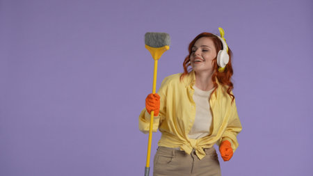 Woman in casual clothing and rubber gloves in headphones holding broom, listening music, smiling. Isolated on purple background.の写真素材