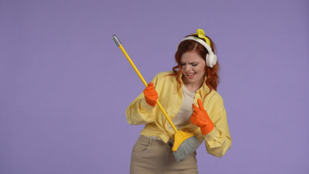 Woman in casual clothing and rubber gloves wearing headphones playing on broom like a guitar enjoying song. Isolated on purple background.の写真素材