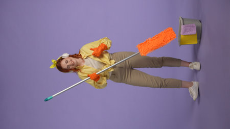 Vertical photo. Woman in casual clothing and rubber gloves in headphones listening music, holding mop, shows thumbs up.の写真素材