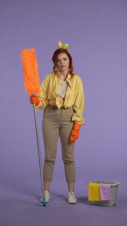 Vertical photo. Woman in casual clothing and rubber gloves holding a mop, tired face expression...