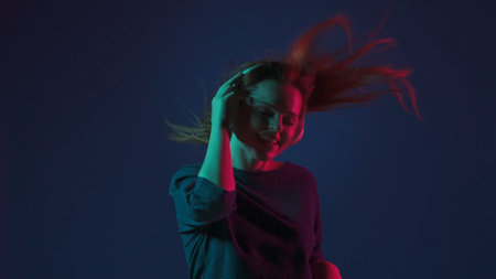 A young redhaired woman wearing white wireless headphones listens to DJ music and dances. Woman in...