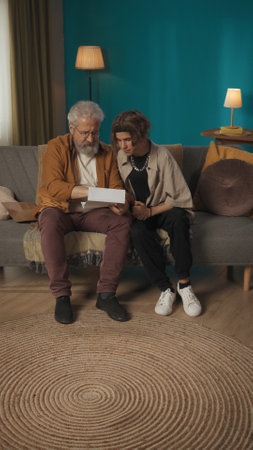 Vertical shot of a teenager, young man sitting with his grandfather on a sofa, couch, reading a...