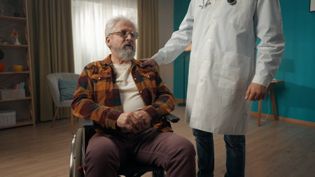 Medium shot of a handicapped eldery patient on a wheelchair having a medical check up at home. The doctor is patting him on the shoulder.の写真素材