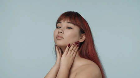 Close-up isolated photo of a young seminude woman with red dyed hair and nude makeup looking at the camera in half turn, running her hands across her neck.の写真素材