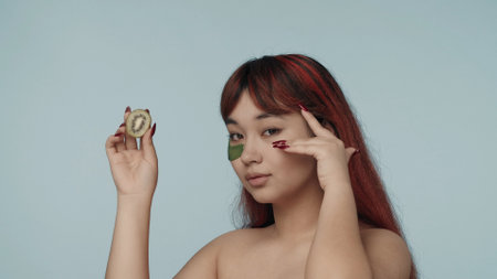 Close-up isolated photo of a young seminude woman with red dyed hair and nude makeup wearing an eyepatch, touching it gently, holding a cut kiwi.の写真素材