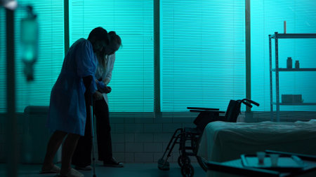 Full-sized silhouette shot a patient with mobility impairment entering the room leaning on a crutch. A female doctor, nurse helps him to get to a wheelchair.の写真素材