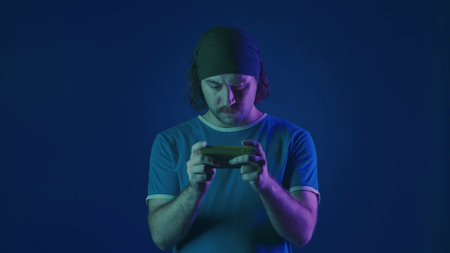 A tense man plays an online game on his smartphone. A man in a studio on a blue background in pink...