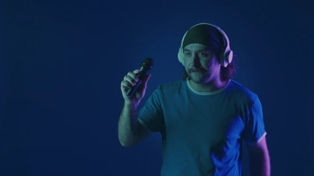 A mustachioed man in white wireless headphones dances and sings into a microphone. Woman in studio on blue background with pink and green neon lights. Karaoke.の写真素材