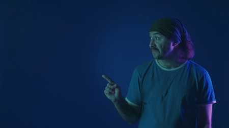 A man points his index finger to the side indicating something. Portrait of a man in a studio on a blue background in pink and green neon light. Advertising area.の写真素材