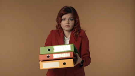 Medium isolated shot of a stressed, anxious and nervous young woman holding a bunch of ring binders in her hands and looking at the camera.の写真素材