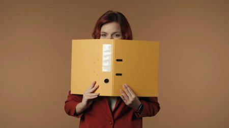 Medium isolated shot of a satisfied, happy and active young woman holding a ring binder in her hands and peeking from behind it.の写真素材