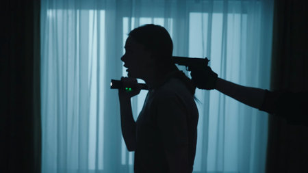 The criminal in black clothes and a balaclava pointed a pistol at the back of the womans head. Frightened woman holding a flashlight held hostage by a burglar in a dark apartment close up.の写真素材
