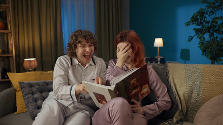 Young couple together. Man and woman in pajamas sitting on the sofa, holding album and looking at old photos joking and smiling.の写真素材