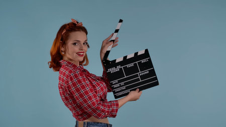 In the frame on a blue background is a young red-haired woman with brightly colored makeup. She looks into the camera and holds a clapper in her hands. Demonstrates filming a movie, TV series HDR BT2020 HLG Material.の写真素材
