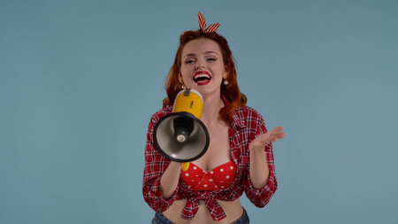 In the picture on a blue background, a young redheaded woman with bright makeup. Looks to the side and speaks through the loudspeaker, the horn. Makes a speech. HDR BT2020 HLG Material.の写真素材