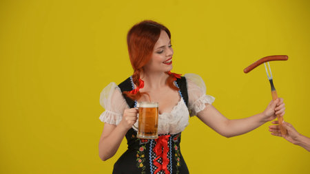 Medium yellow background isolated shot of a young German woman, waitress, wearing a traditional costume, holding a glass of lager, beer and taking, giving a wurst, sausage.の写真素材
