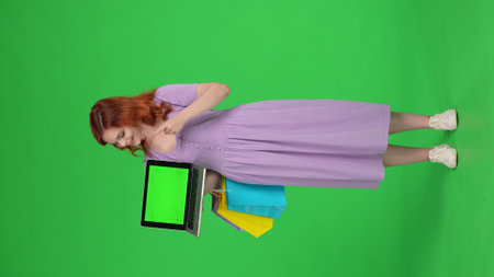 Young woman with shopping bags points her index finger at the green screen of the laptop in her hand and shows thumbs up. Full length woman in studio on green screen. Vertical video. Advertising area.の写真素材