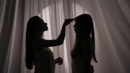 Medium side view shot of two sisters, young womens silhouettes wrapped in a towel. One of them is caressing, brushing the other ones hair in a muffled light.の写真素材