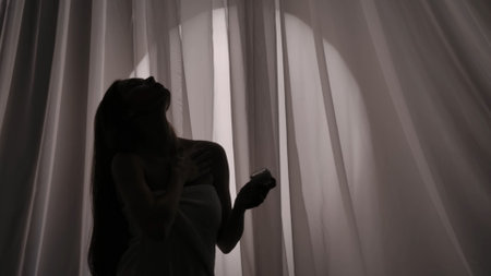 Medium side view shot of a young womans silhouette wrapped in a towel holding a jar of body, face cream and applying the substance on her skin in a muffled light.の写真素材